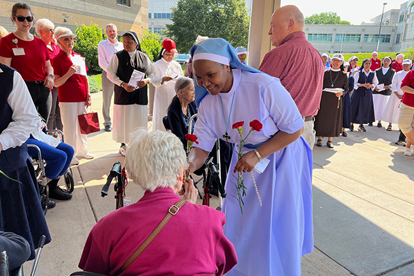 Religious Sister in blue habit hands red flower to lady in pink shirt
